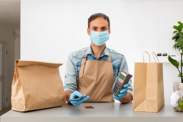 Cashier hand holding credit card reader machine and wearing disposable gloves, paying with smartphone during Covid-19 pandemic.