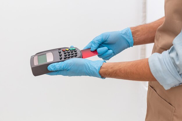 Cashier hand holding credit card reader machine and wearing disposable gloves, paying with smartphone during Covid-19 pandemic.