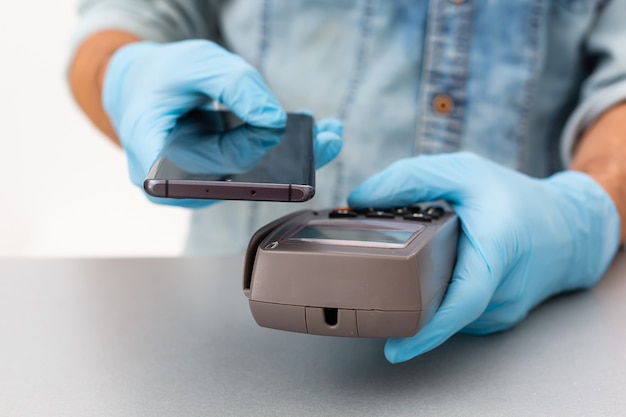 Cashier hand holding credit card reader machine and wearing disposable gloves, paying with smartphone during covid-19 pandemic.