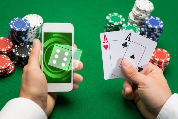 casino, online gambling, technology and people concept - close up of poker player with playing cards, smartphone and chips at green casino table