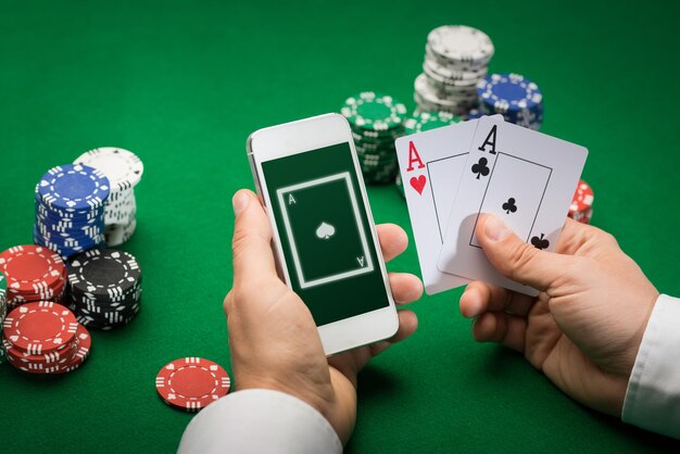 Photo casino, online gambling, technology and people concept - close up of poker player with playing cards, smartphone and chips at green casino table