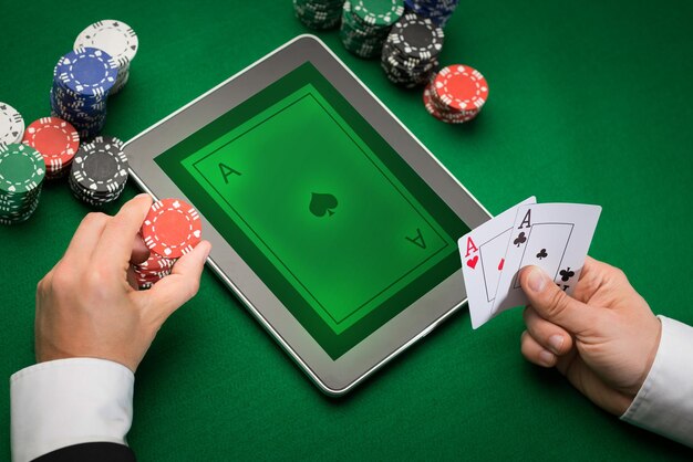 Photo casino, online gambling, technology and people concept - close up of poker player with playing cards, tablet pc computer and chips at green casino table