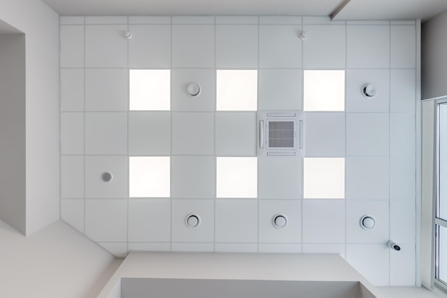 Cassette stretched or suspended ceiling with square halogen\
spots lamps and drywall construction with fire alarm and\
ventilation in empty room in house or office looking up view