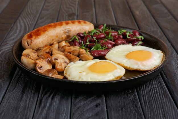 Cast-iron frying pan with fried eggs, sausage, mushroom and beans on wooden table. Low angle view.