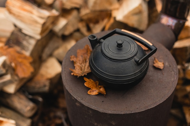 cast iron kettle for traditional oriental tea ceremony on burned stove with beautiful autumn oak leaves and stacked logs on the background