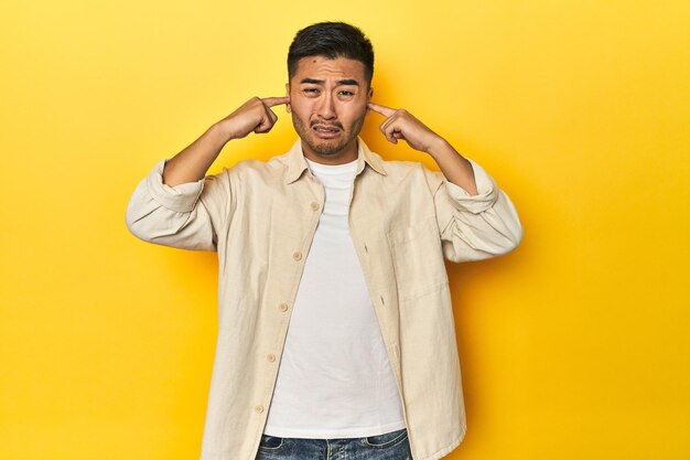 Casual Aziatische man met open shirt wit T-shirt op gele studio bedekt oren met vingers benadrukt