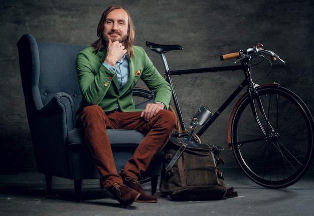 A casual bearded hipster man in a green jacket sits on a chair with single speed bicycle on background.