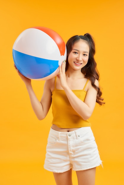 Повседневная красивая азиатская женщина с пляжными аксессуарами, держащая красочный мяч на оранжевом фоне