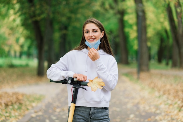 Casual blanke vrouw draagt beschermend gezichtsmasker stedelijke elektrische scooter rijden in stadspark tijdens covid pandemie. Stedelijk mobiliteitsconcept.