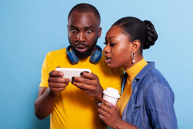 Casual boyfriend and girlfriend watching video on mobile phone, standing over blue background. Modern couple browsing internet together on mobile phone, using social media for entertainment.