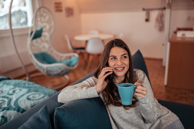 Casual brunette drinking tea and talking on the phone at homely apartment. 