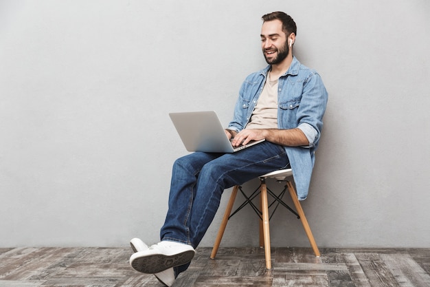 casual brunette man having beard typing laptop and using earpods while sitting on chair isolated over gray wall