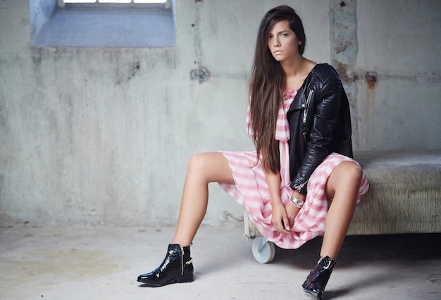 Casual brunette woman in pink dress and black leather jacket sitting on sofa over grey wall.