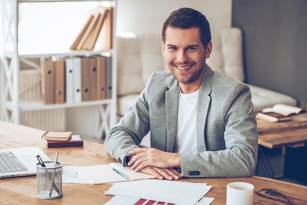 Casual business day. Cheerful handsome young man looking at camera with smile while sitting at his working place