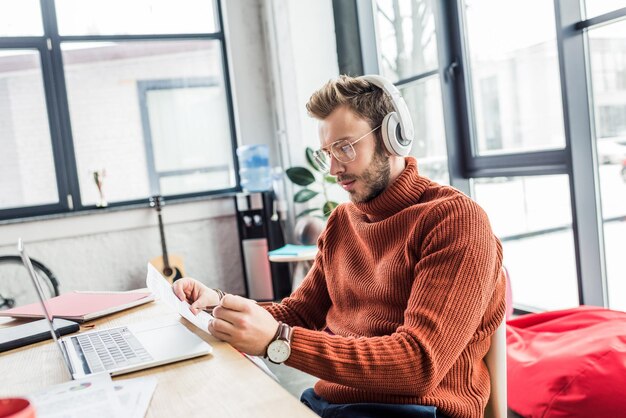 Casual businessman in headphones with document sitting at computer desk in loft office