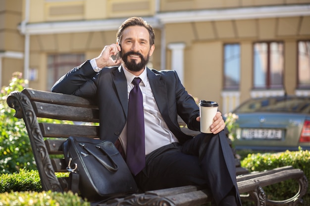 Photo casual conversation. cheerful delighted handsome man talking on phone while drinking coffee