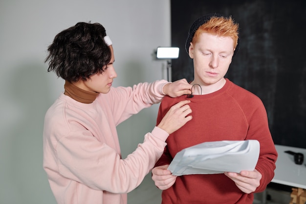 Casual guy with black wavy hair putting microphone on sweatshirt of young male vlogger reading speech on paper