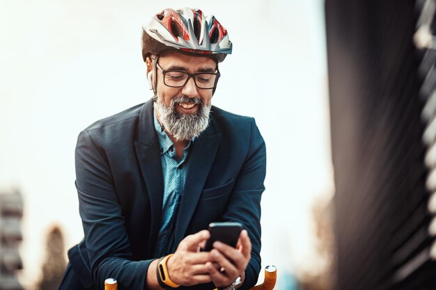 カジュアルでハンサムな中年ビジネスマンが自転車で通勤します。彼は自転車に乗って、オフィス地区の前でスマートフォンを使用しています。