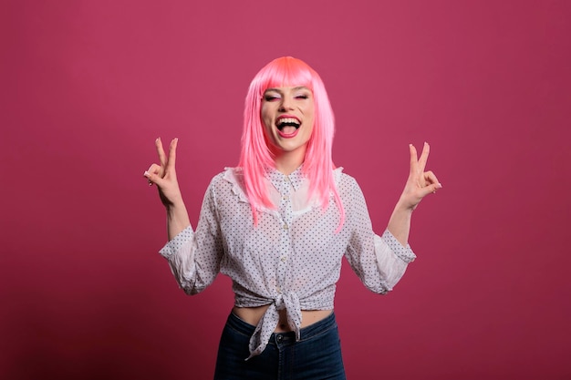 Casual happy person with pink hair doing peace symbol on camera, using two fingers to show victory sign. Feeling positive and optimistic about success, doing peaceful v gesture over background.