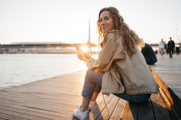 Casual jongedame in jas zittend op een bankje aan de kade van de zee met behulp van smartphone