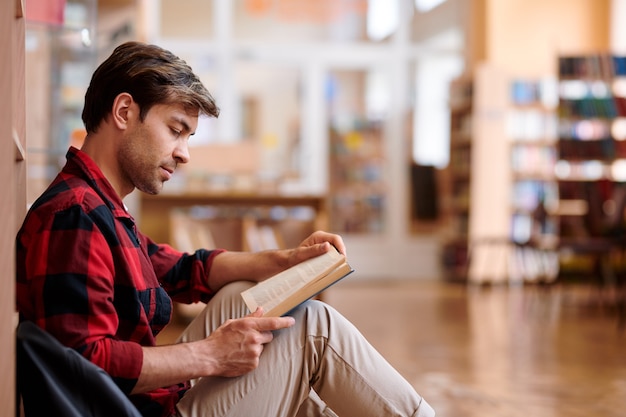 Casual jongeman leesboek zittend door boekenplank in bibliotheek van de universiteit