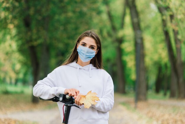 Casual Kaukasisch wijfje dat beschermend gezichtsmasker draagt dat stedelijke elektrische autoped in stadspark berijdt tijdens covid pandemie. Stedelijk mobiliteitsconcept.