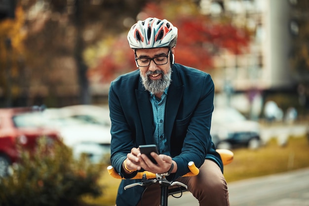 Casual knappe zakenman van middelbare leeftijd gaat met de fiets werken. Hij staat op de fiets en gebruikt een smartphone.