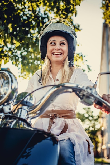 Casual lachende blonde vrouw in moto helm poseren op scooter op natuur achtergrond.
