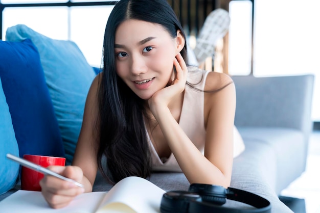 La donna asiatica di stile di vita casual lavora a casa con il taccuino e il caffè sdraiato sul divano con felicità e gioiosa