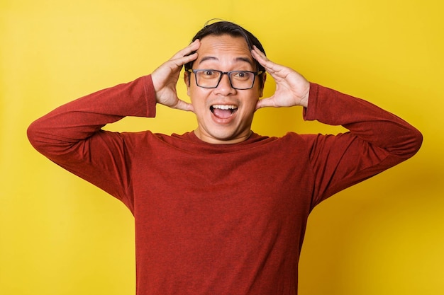 Uomo casual con gli occhiali con espressione sorpresa con le mani che tengono la testa isolata su sfondo giallo