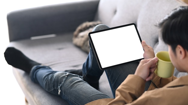 Casual man using digital tablet and drinking coffee while sitting on couch at home.