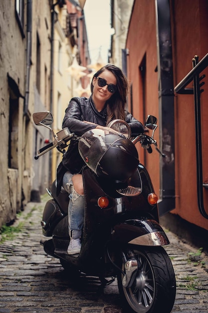 Casual meisje in jeans en leren jas poseren op moto scooter op straat van de oude stad.