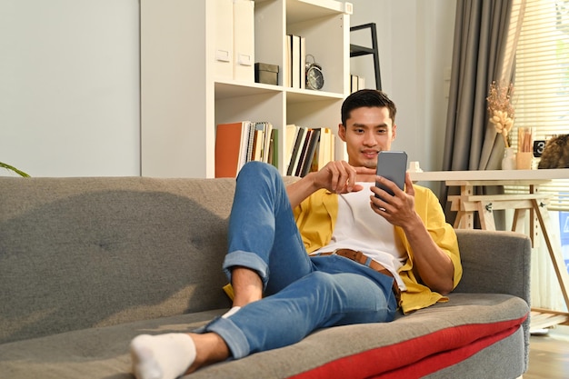 Casual millennial aziatische man ontspannen op de bank thuis sms'en browsen draadloos internet op smartphone