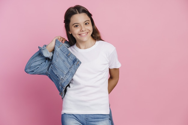 Повседневная, современная девочка-подросток в джинсовой куртке. Девушка искренне улыбается, смотрит в камеру на розовом фоне.