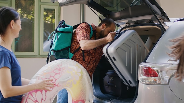 Photo casual mother and father loading luggage into car trunk while daughter bringing beach inflatable. caucasian family getting ready for summer field trip while voyage baggage inside vehicle.