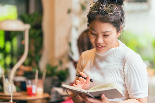 Casual ontspannen aziatische vrouwelijke freelance digitale nomade handschrift schetsboekideeën bekend om haar nieuwe project in café geschoten door coffeeshopraam glasaziatische vrouw zit werk in café vrolijk gevoel