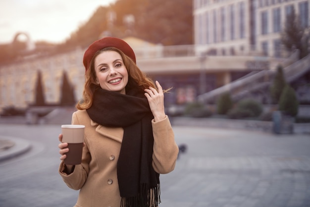 秋のベージュのコートと赤いベレー帽でコーヒーマグを外で保持しているフランス人女性のカジュアルな肖像画