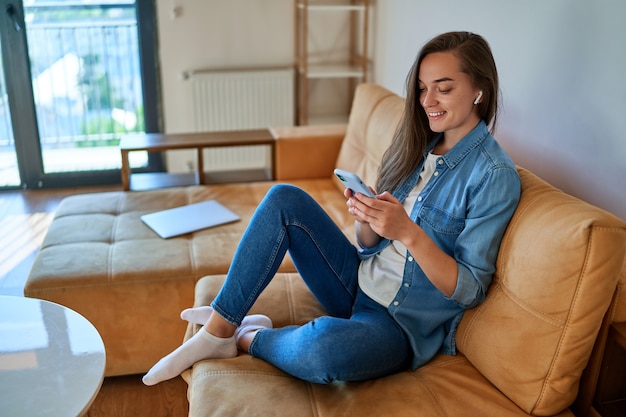 Foto casual slim modern glimlachend millennial meisje met draadloze hoofdtelefoon die smartphone gebruikt voor het bekijken van video-inhoud, het controleren van sociale media en online winkelen tijdens het ontspannen op de bank thuis