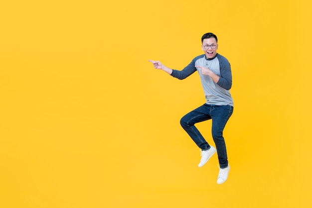 Photo casual smiling asian man jumping and pointing fingers to copy space aside