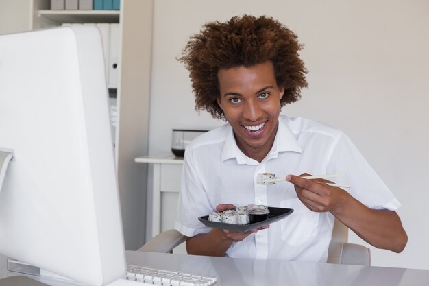 Casual smiling businessman eating sushi at his desk