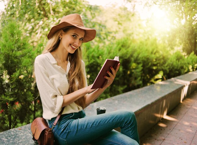 Casual stijl blonde vrouw in vilten hoed lezen van een boek buiten in een stadspark