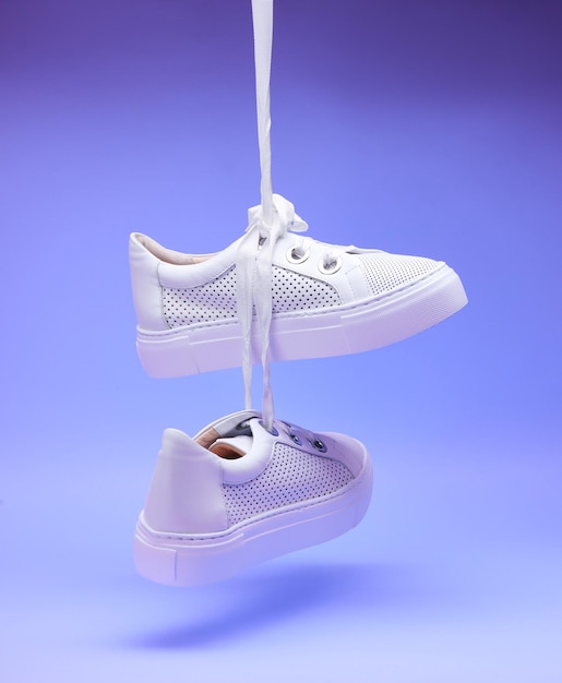 Casual white sneakers hanging on shoelaces in front of gradient purple background perforated leather