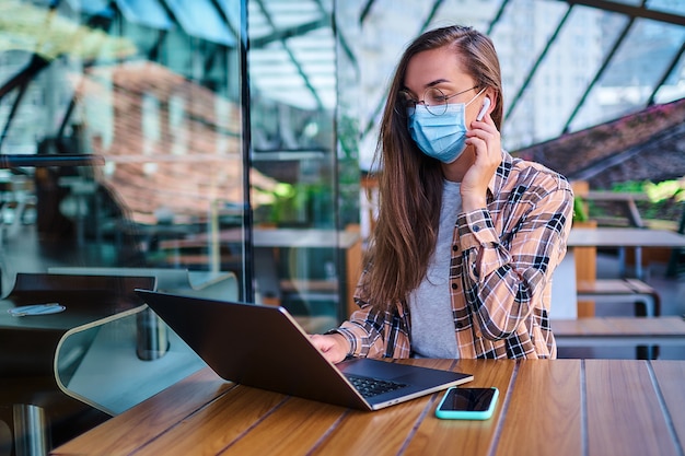 Casual working business woman in face mask, white wireless headphones and eyeglasses remotely works at the computer in cafe. Social distance and health protection in public places