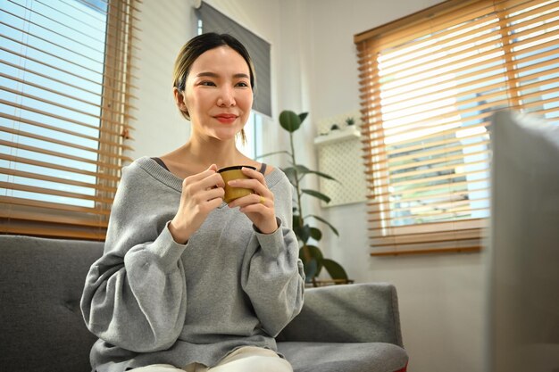 Casual young asian woman sitting on couch drinking coffee and reading online news on laptop at home