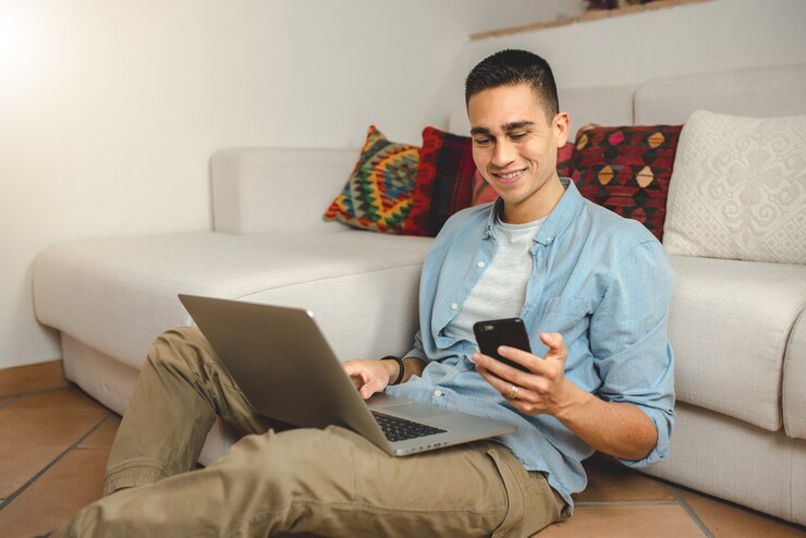 スマートフォンを保持しているコンピューターラップトップに笑みを浮かべて自宅でカジュアルな若い男。