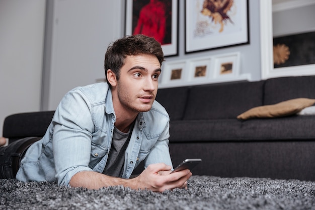 Casual young man lying on a carpet and using mobile phone at home