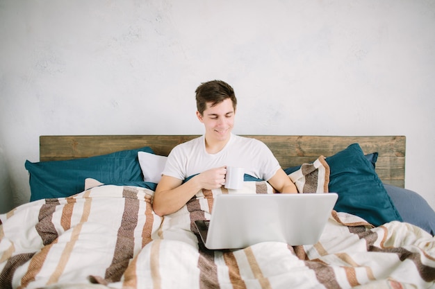 Casual young man using laptop in bed at home and drinking coffee