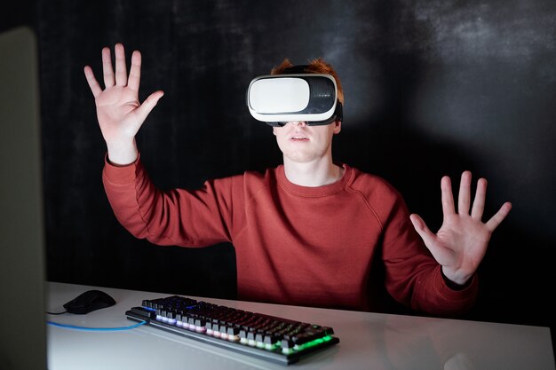 Giovane casual in cuffie vr seduto da scrivania davanti allo schermo del computer virtuale nell'oscurità