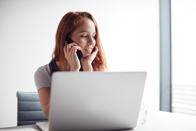 Photo casually dressed young businesswoman working on laptop at desk in modern workplace making phone call