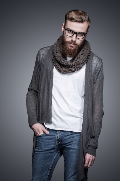 Casually handsome. Handsome young bearded man in eyeglasses holding one hand in pocket and looking at camera while standing against grey background